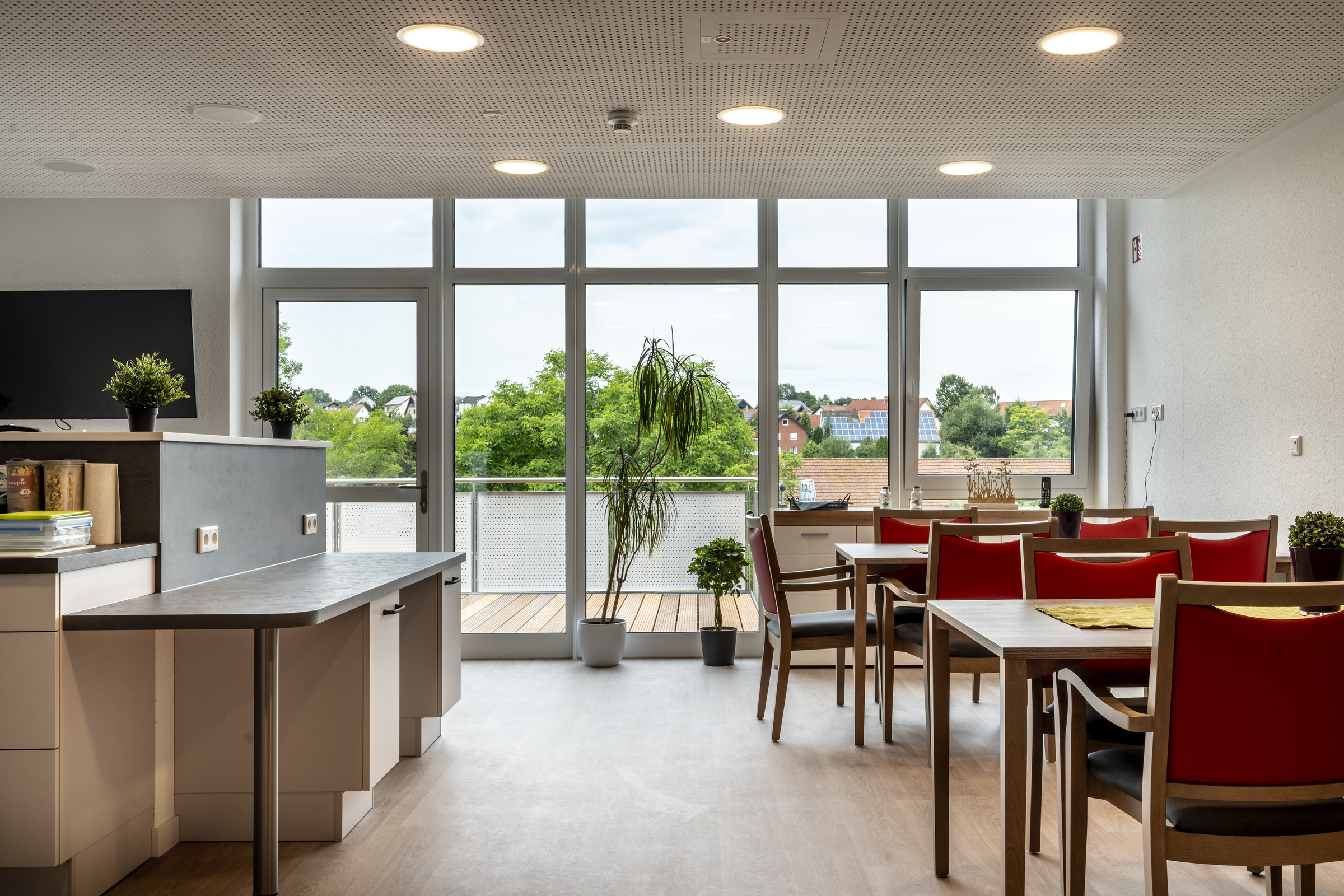 Gemeinschaftsraum im Meierhof mit Küchenzeile, Esstischen mit roten Stühlen und großer Fensterfront zur Terrasse