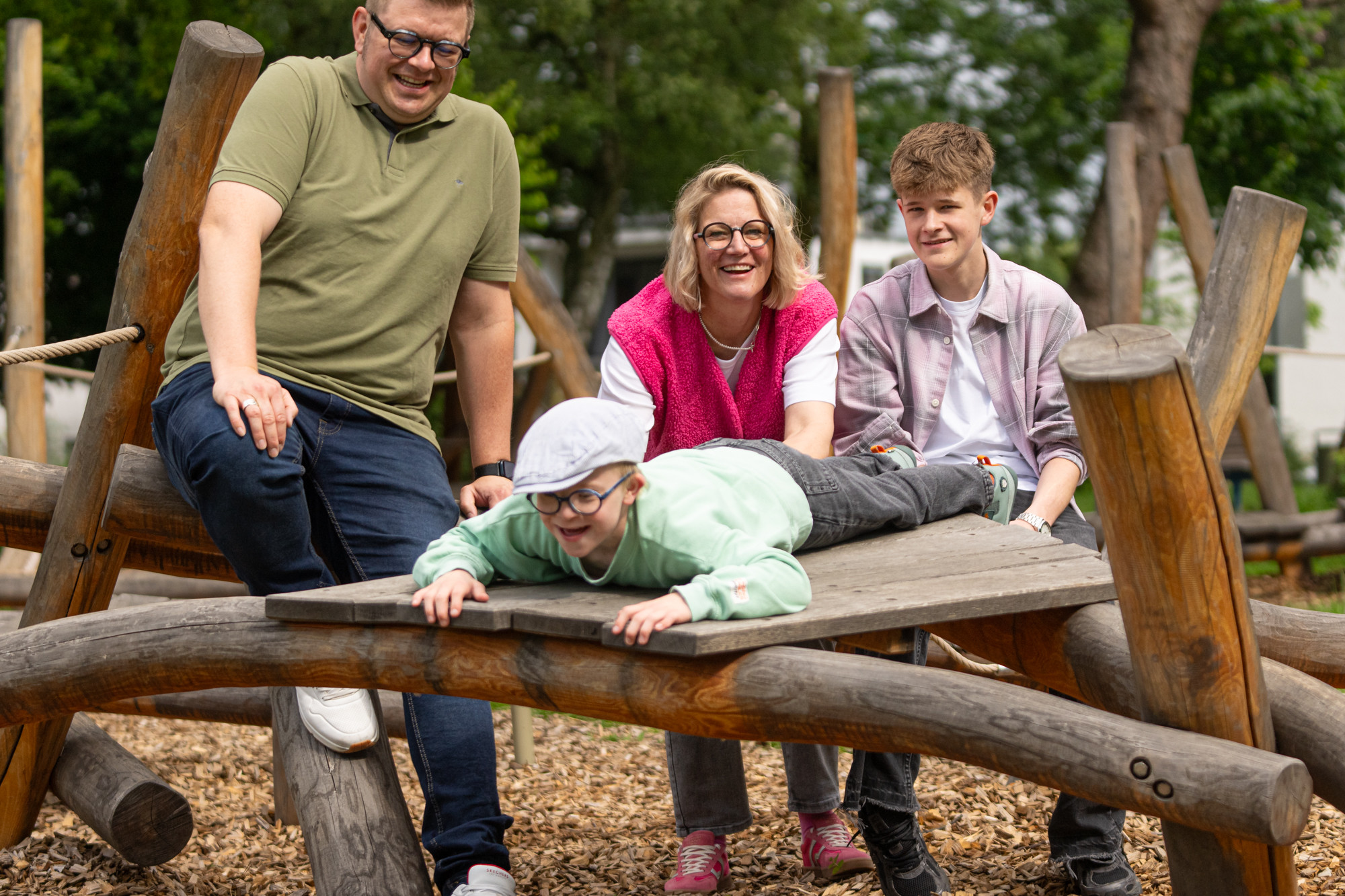 Eine Familie auf dem Spielplatz: Eltern und zwei Kinder lachen gemeinsam an einem Klettergerüst aus Holz